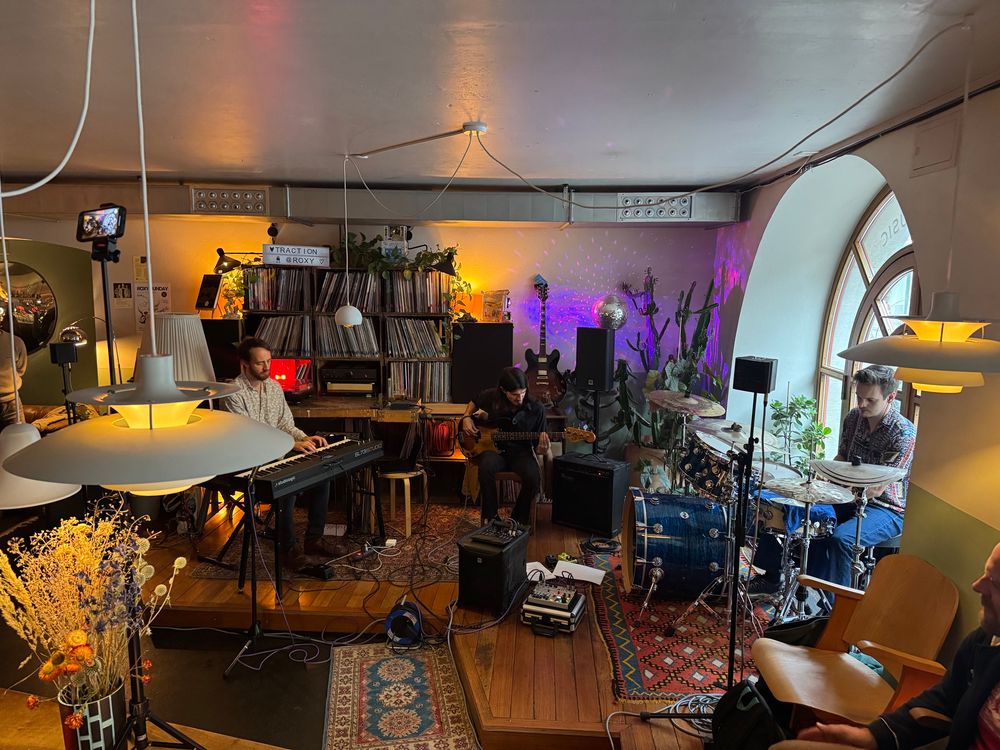 A jazz trio on stage in a small vinyl cafe with lights and flowers in the foreground. 