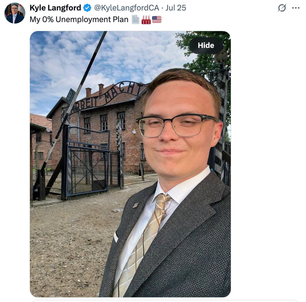 Screenshot of a tweet by Kyle Langford, dated July 25, showing him smirking in a suit in front of the Auschwitz death camp gate. The tweet reads: “My 0% Unemployment Plan” with emojis of paper, a factory, and an American flag. The infamous “Arbeit Macht Frei” sign is visible behind him.