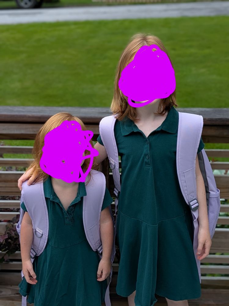 Two kids in matching green dresses and purple backpacks on their way to school