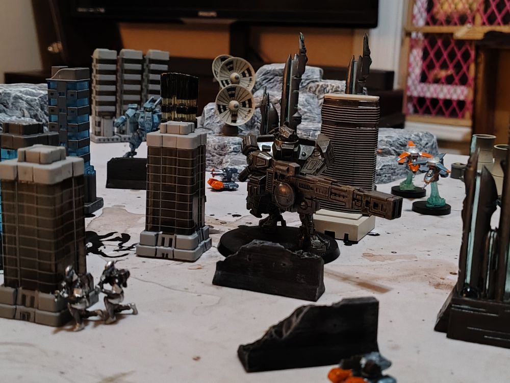 Tiny plastic robots with tiny plastic buildings, near some tiny plastic infantry surrounded by tiny plastic vehicles.