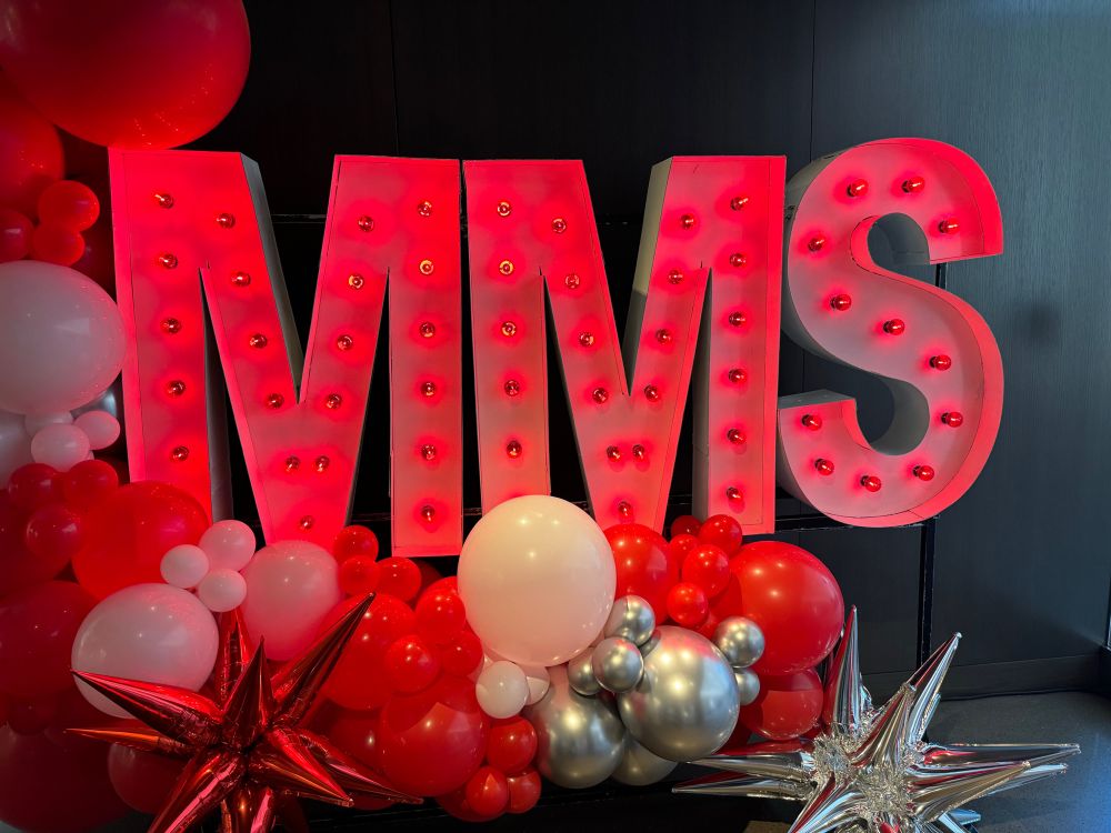 A sign with the letters MMS in red surrounded by balloons and decorations. 