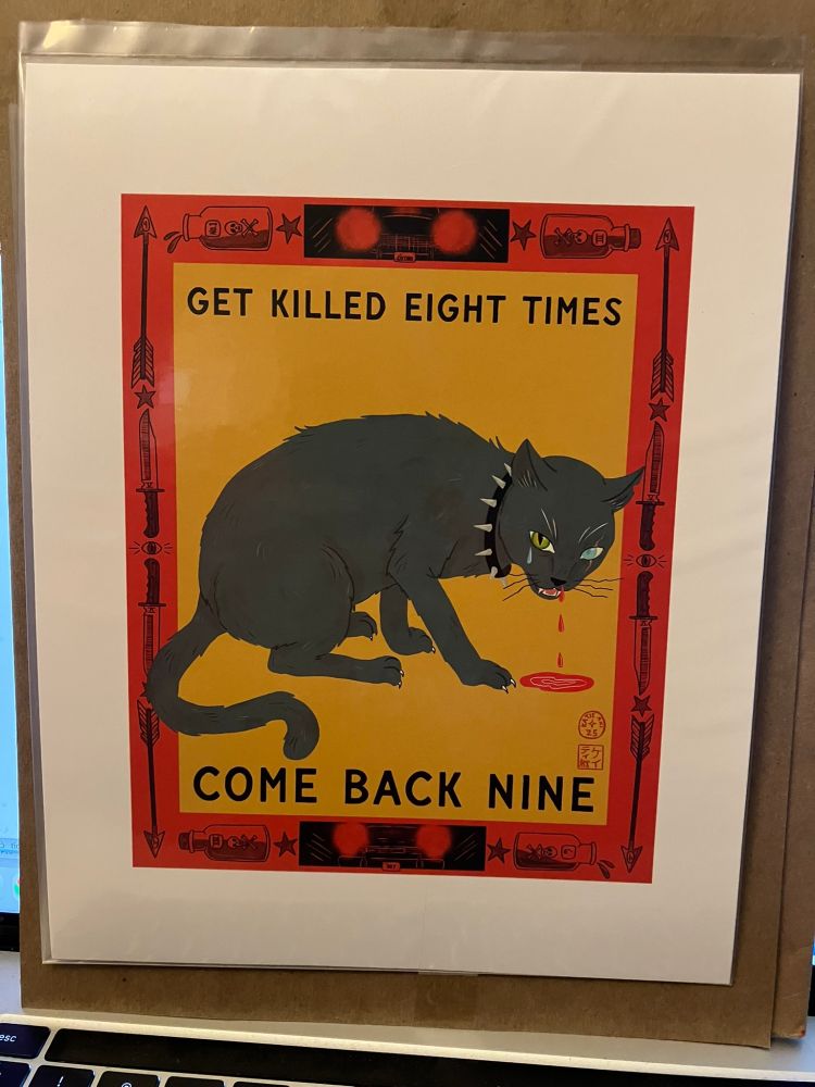 shows a print in plastic leaning against a laptop screen.  the pring is the grey cat print below which reads "get killed eight times come back nine"