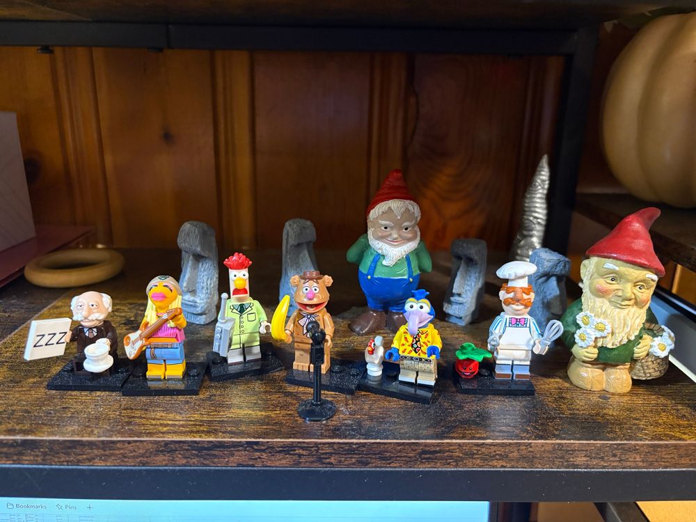 A bunch of muppet lego figures, a couple of gnomes, and some Easter island heads on a brown shelf