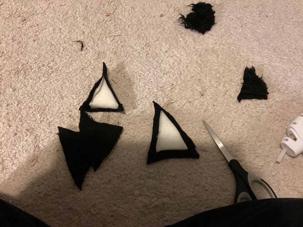 Two triangular pieces of foam on pieces of black fabric. There is also a pair of scissors, fluffy pieces of fabric, and a glue gun. They are all laying on a light gray carpet.
