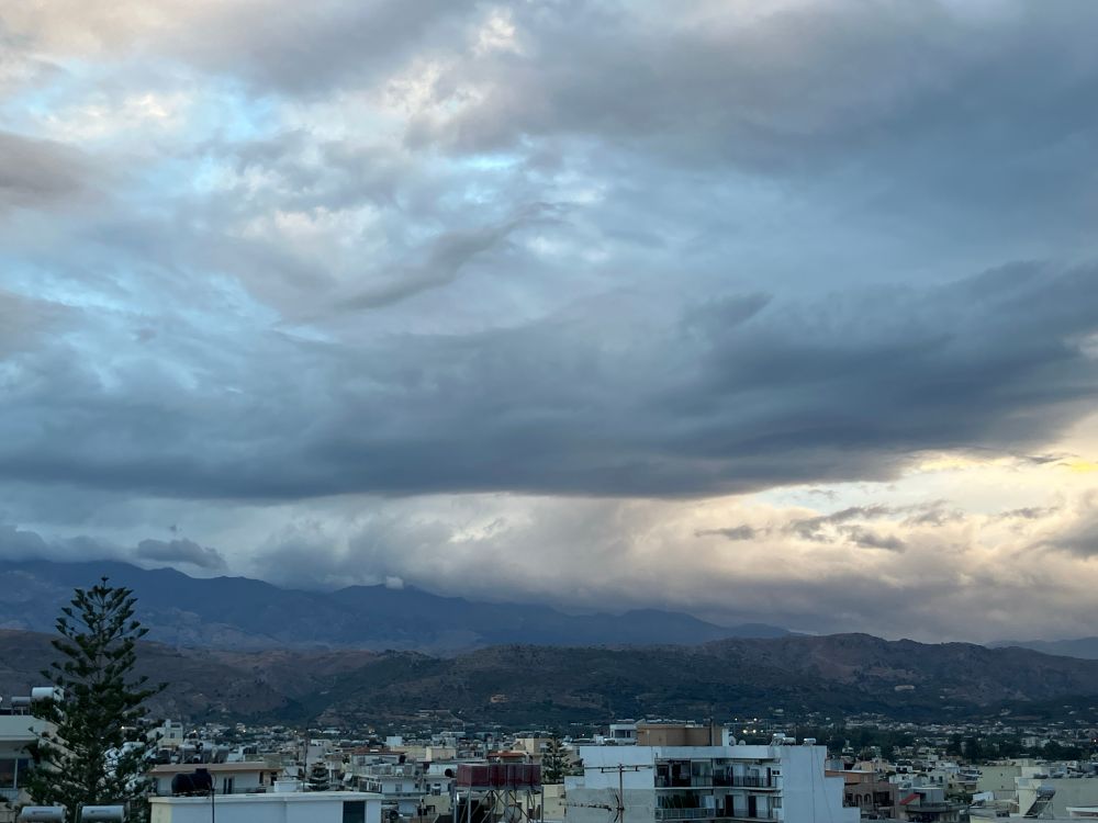 Rainy looking clouds at sunset over a city skyline, behind which is a dark mountain range