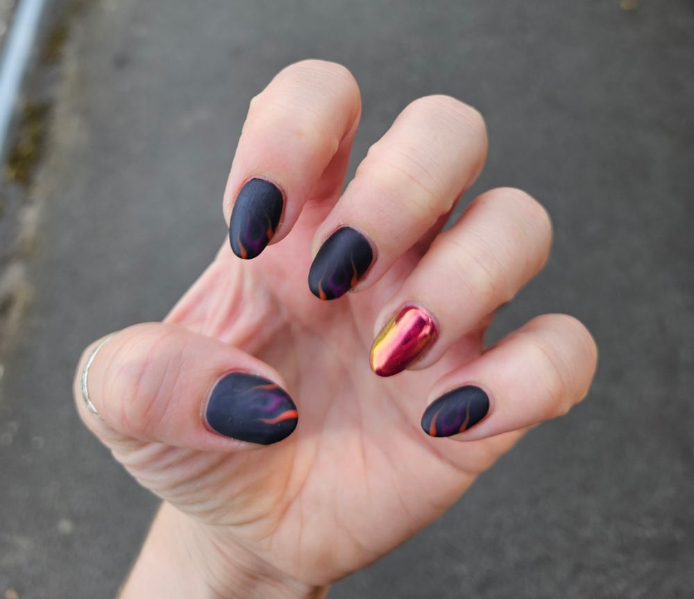 Close up of my left hand, fingers curled in. All of the nails except the ring finger are black with orange and purple flame designs on the side. The ring finger is bright pink with a gold shimmer over the top.