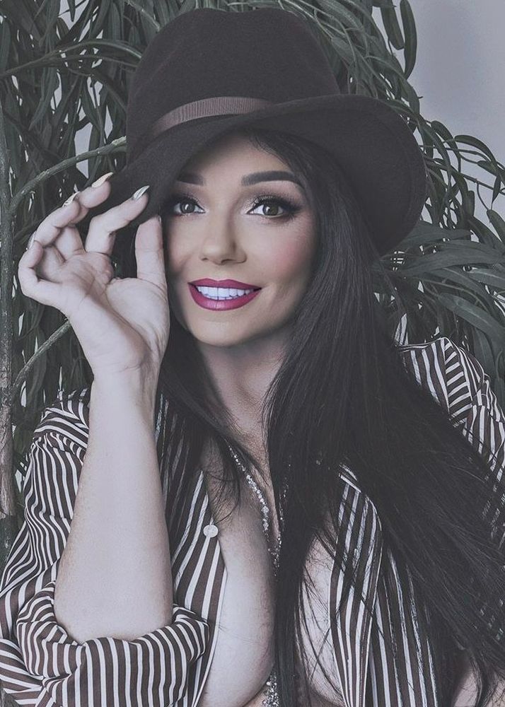 Charlene with long dark hair playfully posing while holding the brim of a stylish black hat. She is wearing a striped shirt that is unbuttoned, with a captivating smile and bold red lipstick. The background features lush green foliage, adding a natural touch to the composition.