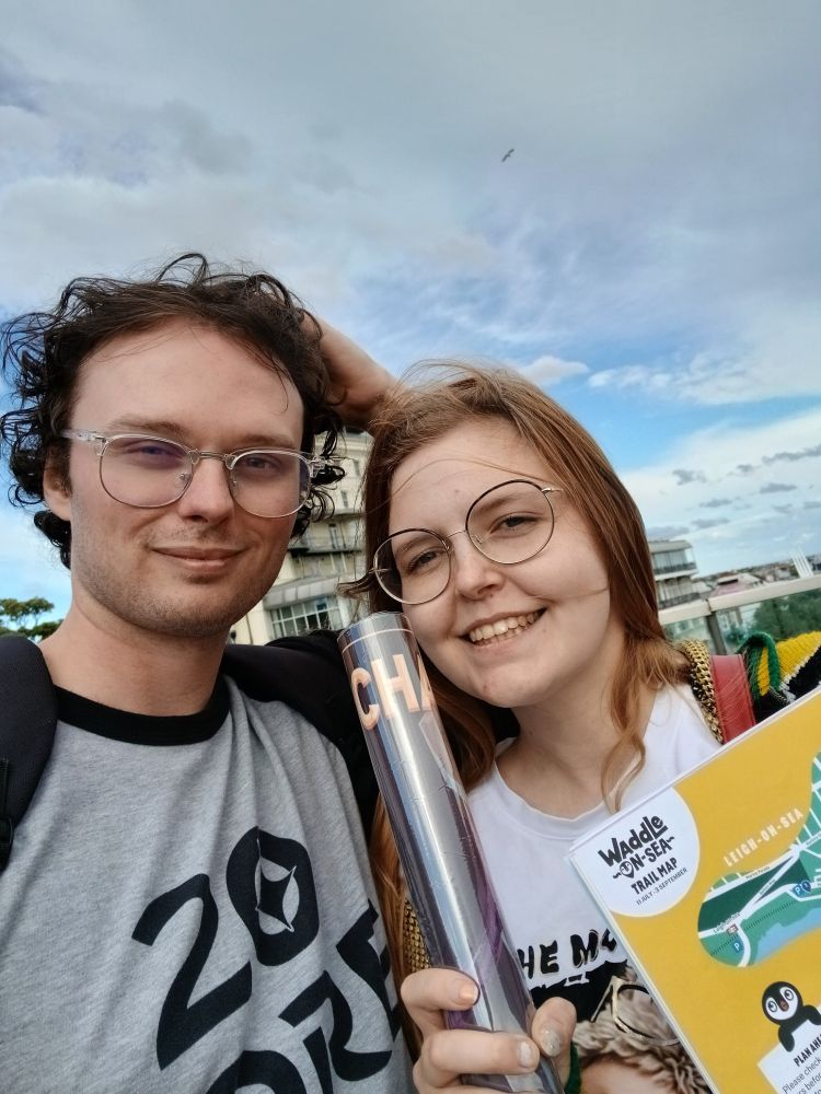 A selfie of Adam and Allie smiling on Southend Pier holding the jafar set champion mat and a waddle on sea sticker book
