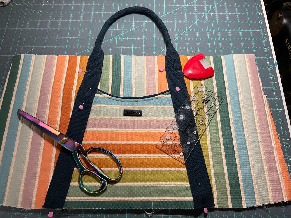 The front of a tote bag in progress sits on my work table. The fabric is wide stripes of different summery colours on cream, with navy webbing for handles. The stripes go vertically on the body of the bag, and horizontally on what will be the front pocket. The pocket is topped with navy piping, and the handle webbing is pinned to the bag fabric with pink pins. On the pocket is a gunmetal label reading “handmade” with the outline of a bird. 