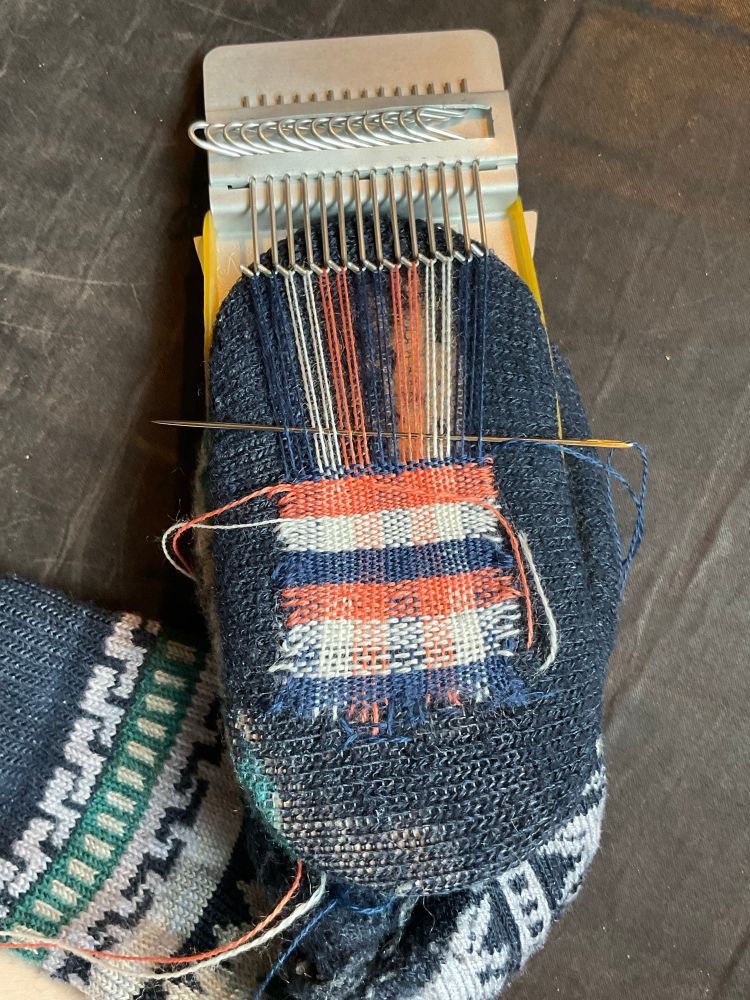 The navy sole of a sock is stretched across a mending loom (a wooden disc and metal plate with hooks that flip back and forth.) The weaving in progress is a plaid of blue, white, and orange. 

Loom by AlexonVer on Etsy; thread and very long darning needle by Sue Spargo. 