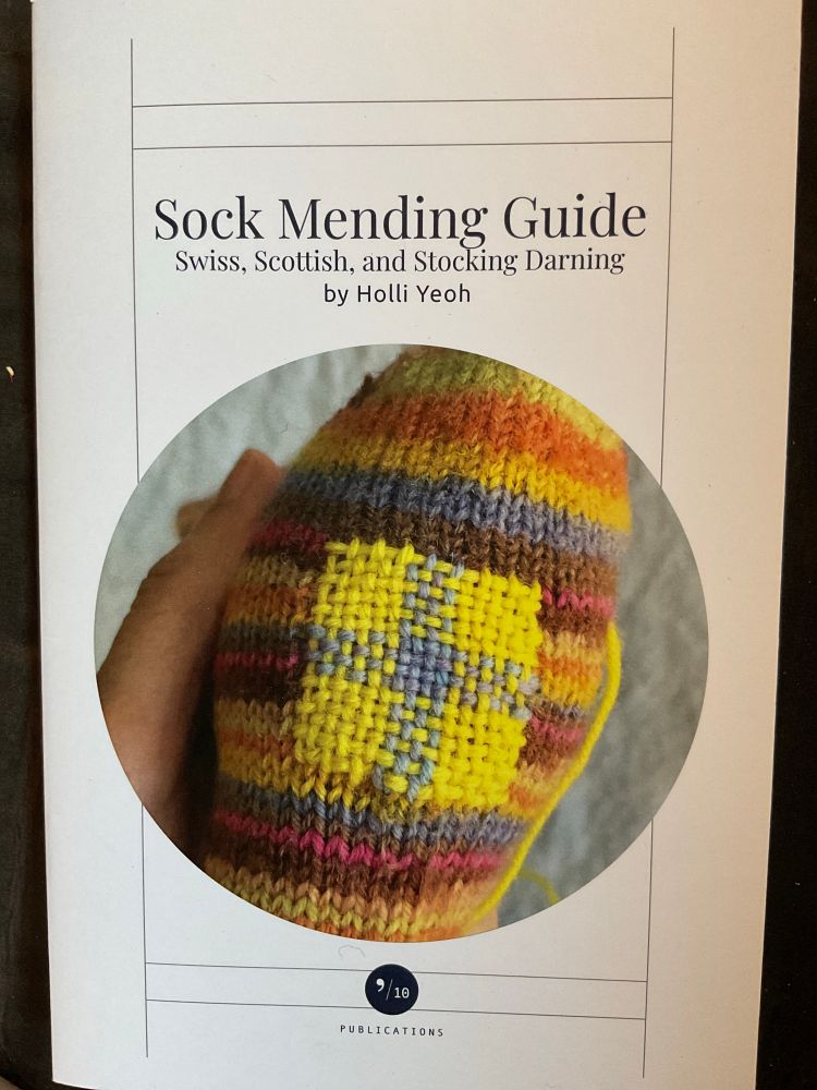 Booklet cover: Sock Mending Guide by Holli Yeoh

White paper with black text, and a photo of a pale hand holding up a striped sock with a blue and yellow woven patch. 