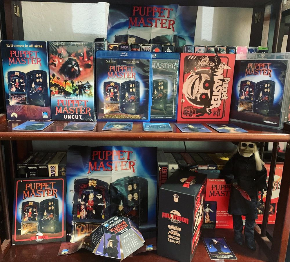 Collection photo related to the original Puppet Master. Top shelf features various copies of Puppet Master: VHS, Uncut VHS, first release Blu-ray, Blu-ray from the trunk box set, Umbrella lobby card style slipcover, Umbrella Blu-ray, plus original holographic promotional trading cards from 1989. 
Bottom shelf: standee, fold-out poster, 1989 promotional calendar, Full Moon Fright Box 2 box set, sleeve for the promo calendar, Tunneler and Leech Woman trading cards from the action figure series featuring '89 promo stills, and 12" Blade doll.