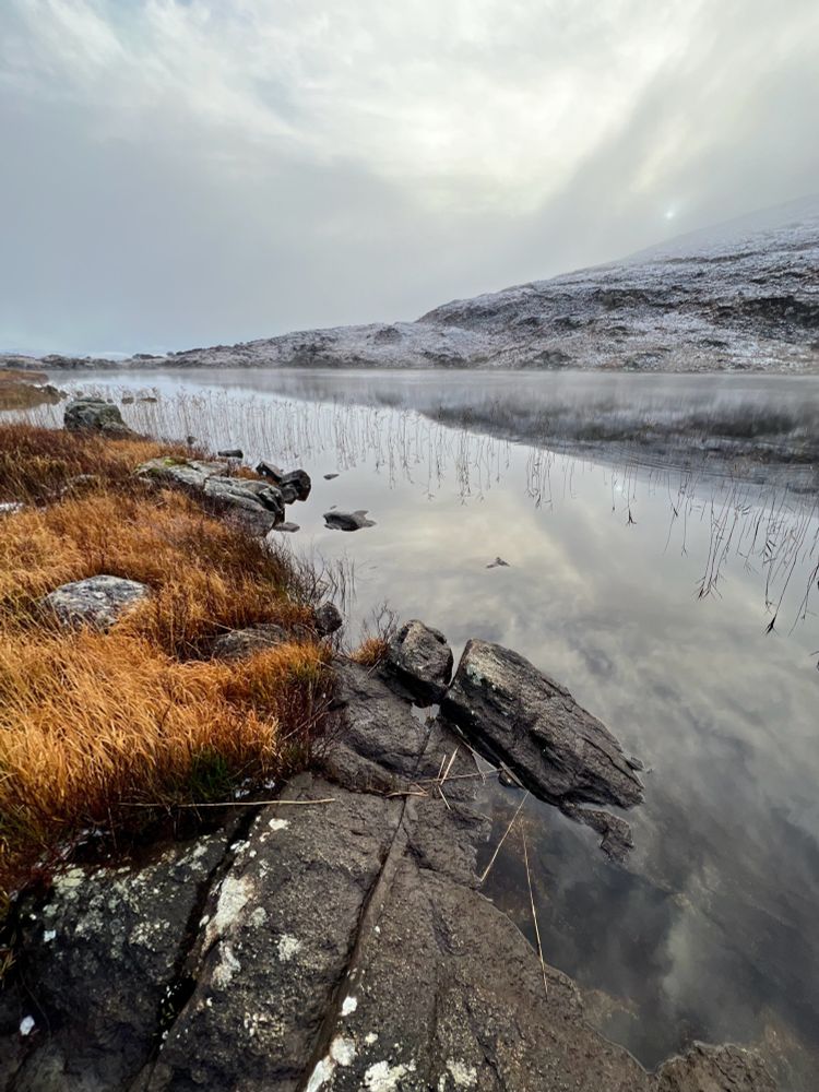 Snow on a hill beside a lake with dramatic reflections of grey clouds.