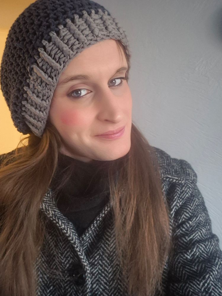 A picture of Lavender wearing a dark blue and Grey knitted beanie. Her reddish brown hair is split on either sides down about chest height. Shes wearing a black and white herringbone coat and a black scarf underneath. She is smiling and has light pink lip gloss on.