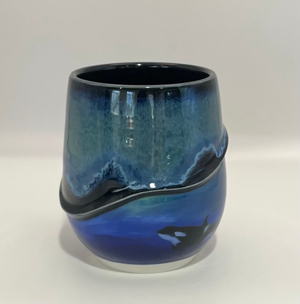 This view shows the same mug that has a relief ribbon separating the top portion from the bottom portion. The ribbon has swooped to a much lower position on this side of the mug. The top portion is a flowing blend of black, blue and green glaze colors. However, from this view one can only really see the orca calf swimming through blue waters on the bottom portion.