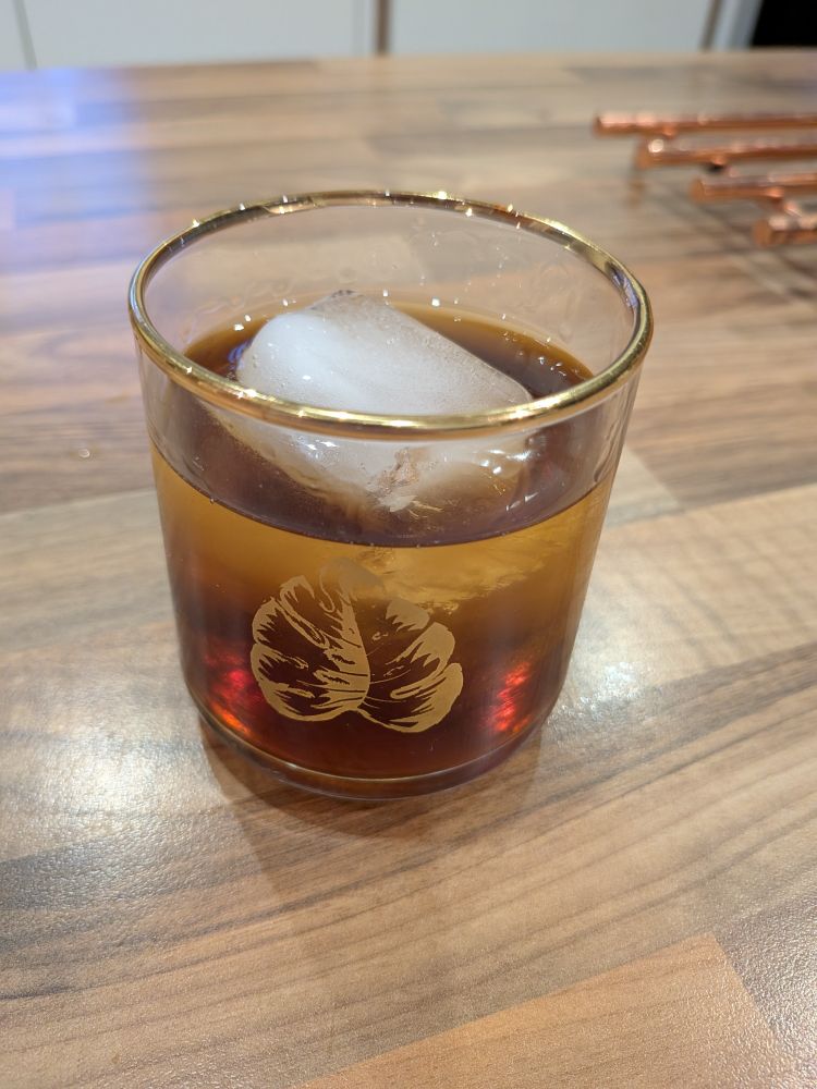 A Hazelnut Old-Fashioned cocktail in a good rimmed glass with a gold leaf on the front. The cocktail has a giant ice cube in it and stands on a brown wood-effect kitchen surface.