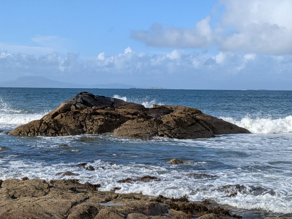 Waves breaking against a rock. The sky behind is mostly blue with a few clouds