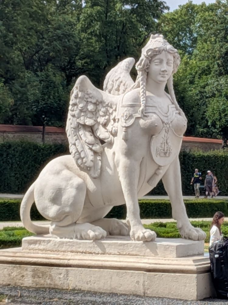 A statue of a mythical creature with the torso and head of a woman, the hindquarters of a lion, and wings. The statue is looking directly towards the camera with a smug expression.