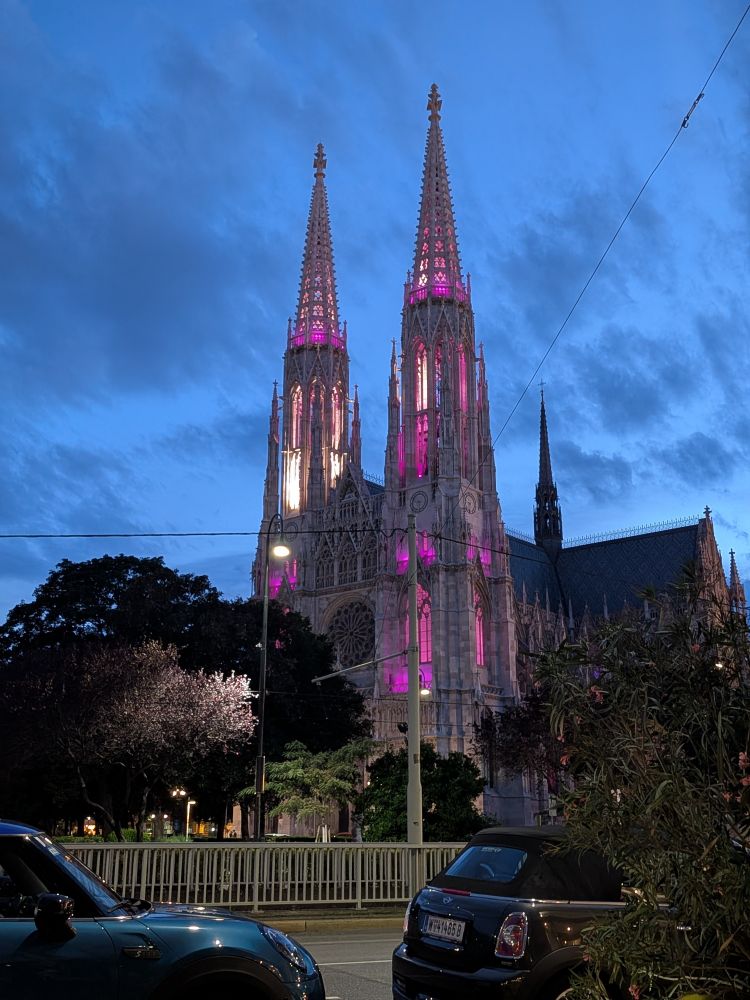 The twin spires of Vienna's Votivkirche rise against a sky of darkening blue and mottled grey cloud. They are lit internally with a combination of purple, pink and white light.