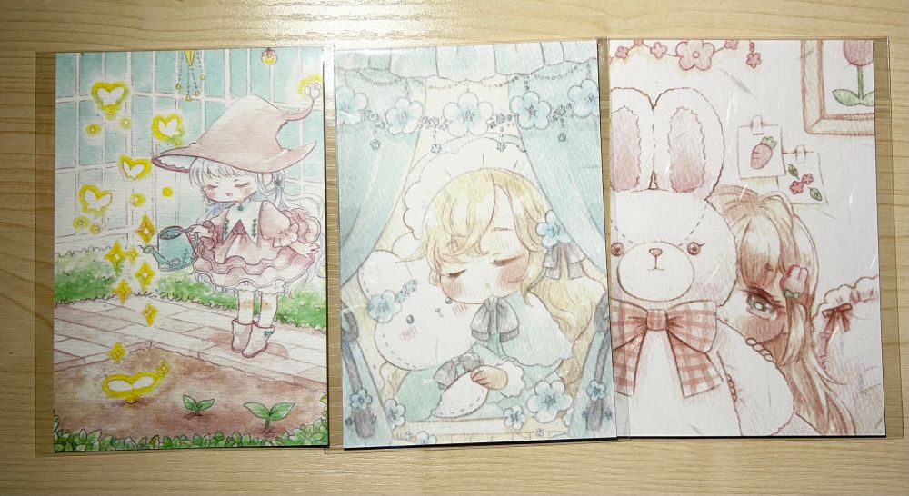 ポストカード3点を映した写真。左から少女が小さな芽を魔法で育てている煌びやかなイラスト。白いウサギのぬいぐるみを目を閉じて静かに抱き抱える少女のイラスト。大きなウサギのぬいぐるみの影に隠れて恥ずかしがっている少女のイラスト。