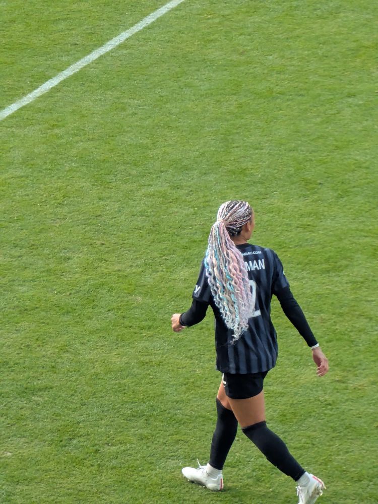 Trinity Rodman with pink and blue hair over a blue and black jersey on a soccer field