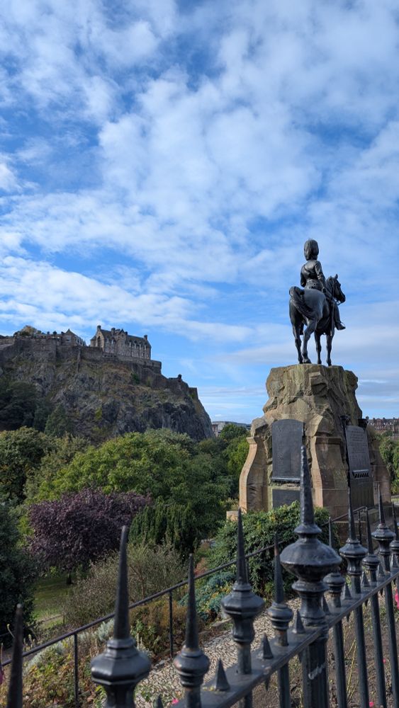 Das Edinburgh Castle auf einem Hügel. Der Himmel ist bewölkt. Im Vordergrund eine Statue mit einem Mann auf einem Pferd und davor ein Zaun. 