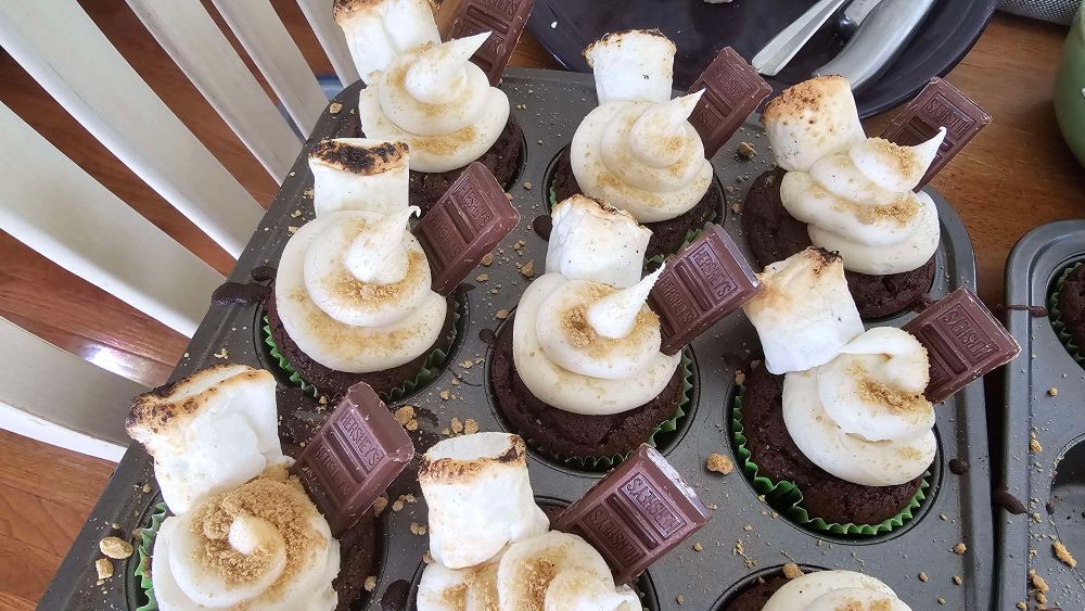 s'mores cupcakes in a pan. Chocolate cupcakes with marshmallow cream frosting, topped with graham cracker crumbs. Each one has a toasted marshmallow and a bar of Hershey's chocolate pressed into the frosting.
