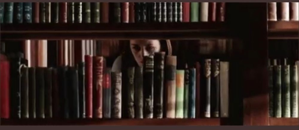 Das Gesicht einer Frau hinter einem offenen Bücherregal einer Bibliothek. Sie schaut suchend in die mit Büchern gefüllten Regale.