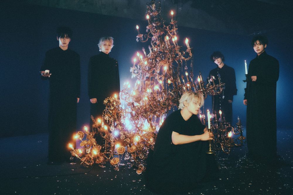 all 5 txt members in all-black outfits with a chandelier that's bigger than any of the members by like a lot. the vibes are great, soft lighting on a dark background, very cool. some of them are holding candles which i think is nice.