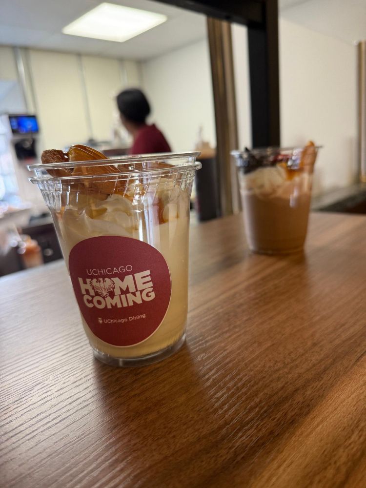A milkshake with a "UChicago Homecoming" label on the side, placed on a wooden table. In the background, an individual is working at a counter and there are two beverage cups.