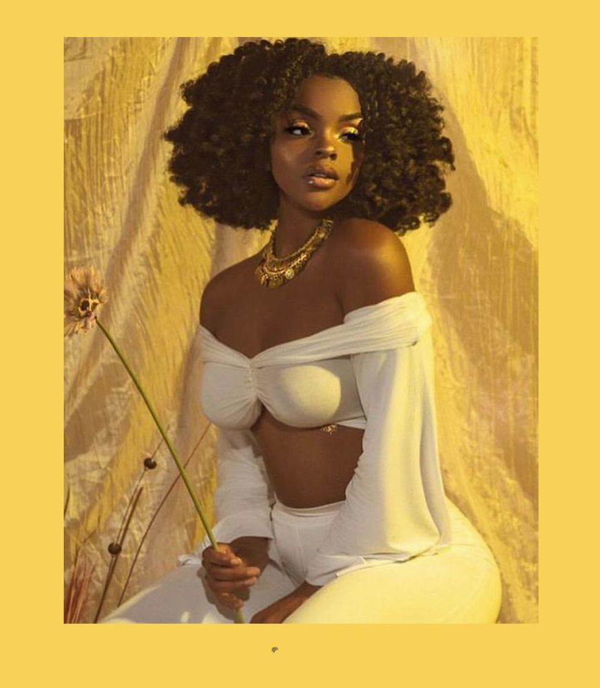 Photograph of a seated black woman wearing a white top with long flowing sleeves and white pants. She is wearing a gold necklace and looking to the right side of the image.