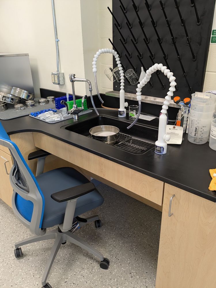 Accessible Sink is shown with chair in the knee space for scale. The sink has two white DI water faucets, as well as a tap water faucet and an eye wash. To one side of the sink is a drying station with numerous sieves and falcon tubes. To the other side of the sink are some empty beakers and DI water wash bottles. There is a beaker drying rack on the wall behind the sink, and a 8" washing sieve in the sink for scale 