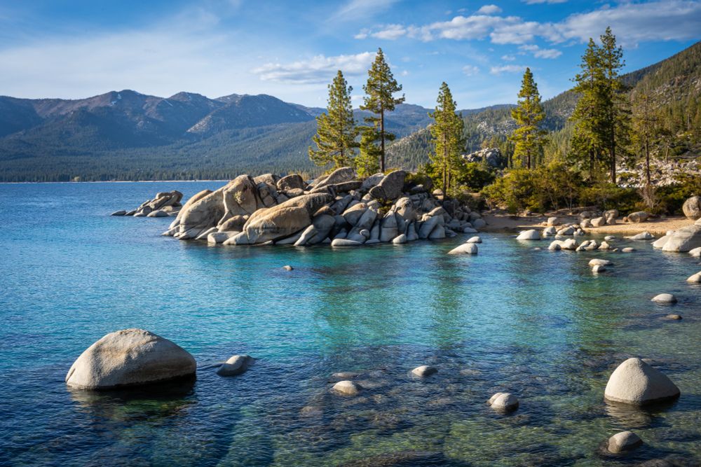 Crystal clear blue water fills most of the frame, with a few rounded rocks peaking through the surface. In the center of the photo is an outcropping of boulders with pine trees, while in the distances are green rolling mountains under a blue sky.