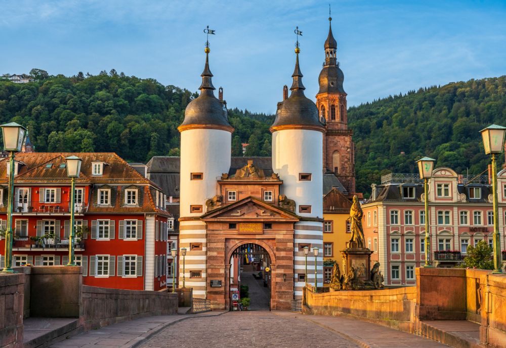 Two baroque towers sandwich a stone gate at the edge of a cobbled bridge. In the background is a church spire and the medieval old town of Heidelberg. 