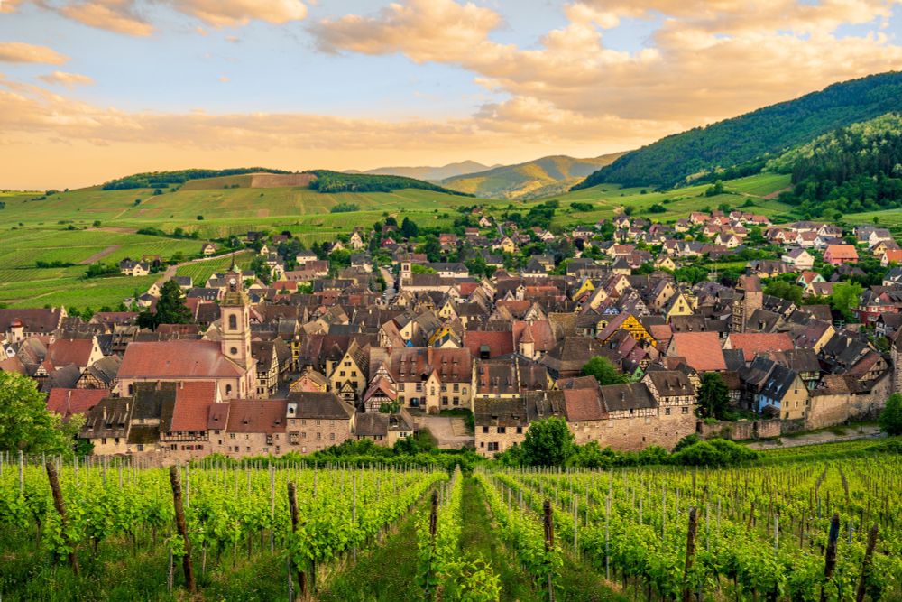 A small French village is nestled in a bowl of green vineyards under an orange clouded sky. 