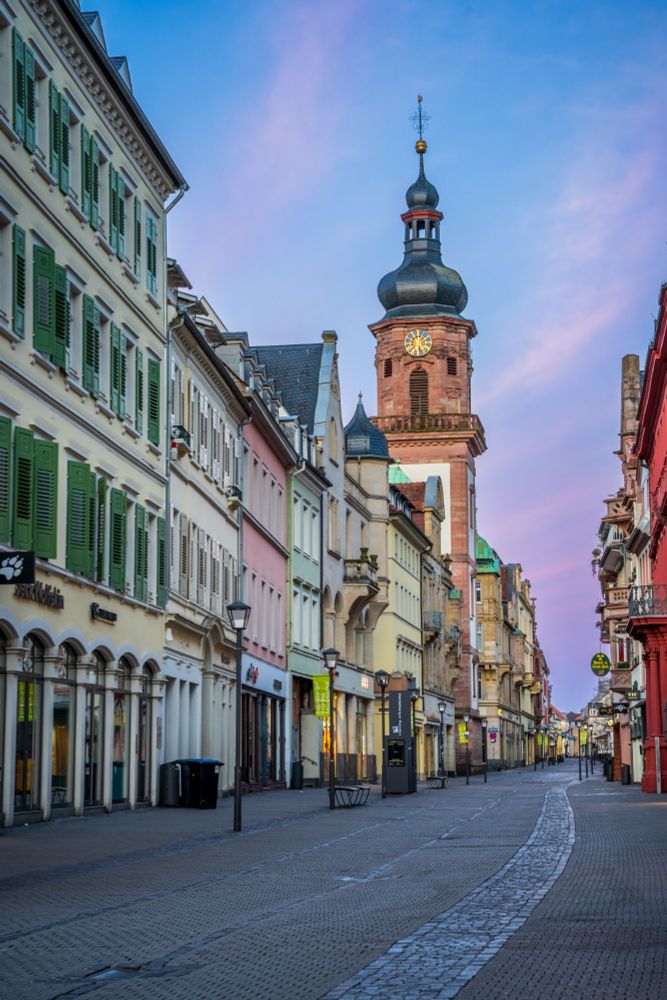 The cobbled walking street of Heidelberg, Germany. Pastel houses dating back hundreds of years line the streets while the domed spire of a Baroque church reach into a pink sky.