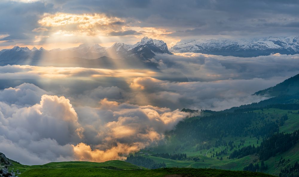 A valley fills with rolling clouds, covering green hills. In the distance are snow capped mountains, while a single whole in the clouds above let in a powerful beam of golden light.