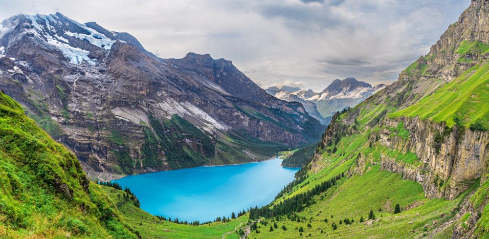 A vista view of a high altitude valley in the Alps. In the foreground, the sloping walls of the mountains are covered in luscious green grass, leading down to a glacial lake. In distances are rocky mountains topped with glaciers.