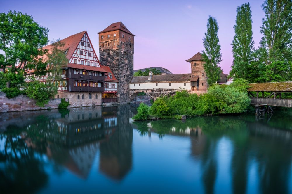A glassy blue river fills the foreground. A wooden bridge extends from the right of the frame to an island with a tower. On the other side of the island a stone bridge leads to an even taller tower and timber framed house.