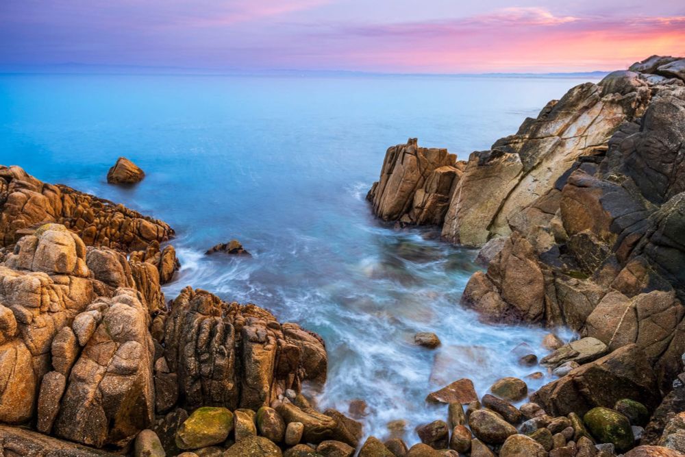 A light blue ocean is hazy and swirls with white lines. Around it are bright rocky cliffs being lit by a cotton candy sky.