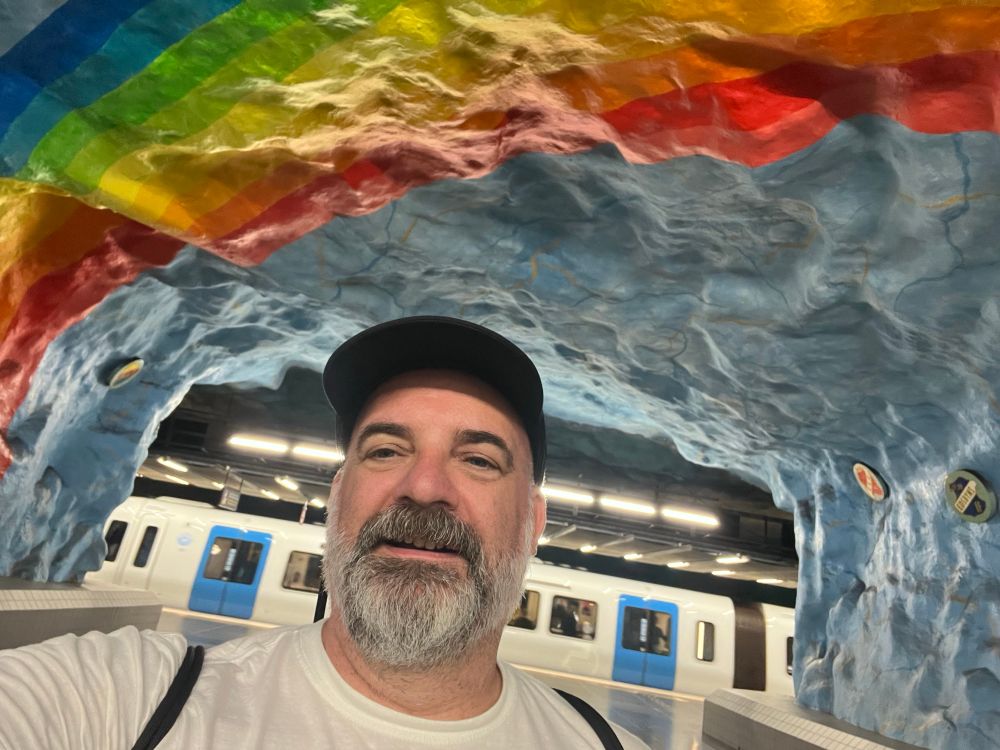 Me, a man with a rapidly greying beard in a metro station with a rainbow painted on the cave-like roof