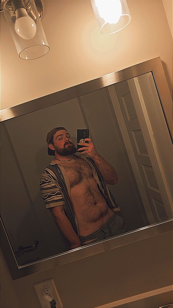 Mirror selfie of a man with beard, wearing a ball cap and an open button-up shirt. Hairy chest and stomach, fit build but a small tummy no abs. 