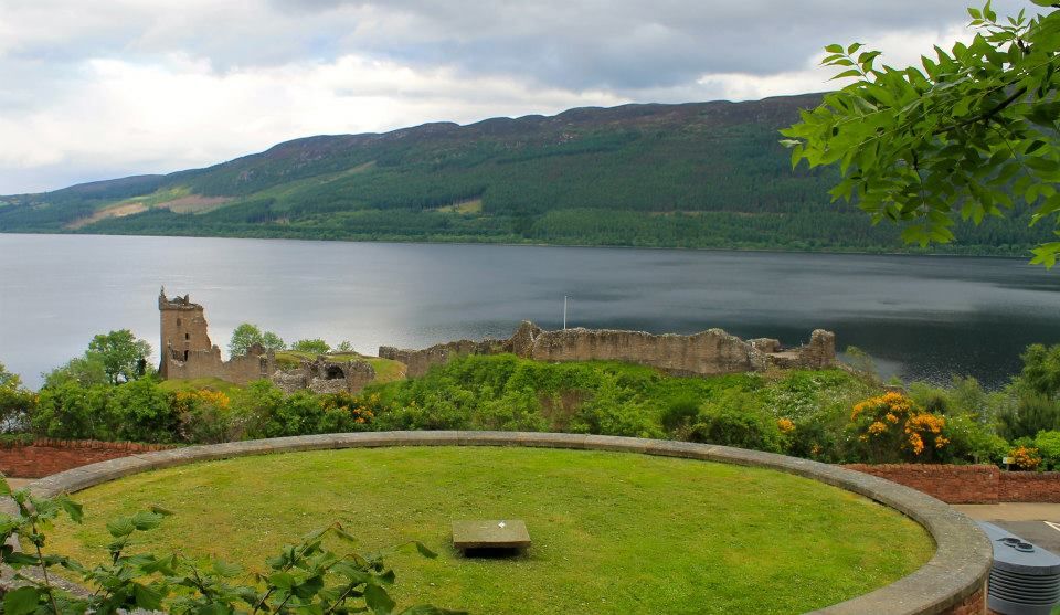 The ruins from a different angle. In the foreground is a building, of which only the roof is visible; this is a green grassy plain. In the background are water and a hilly landscape.