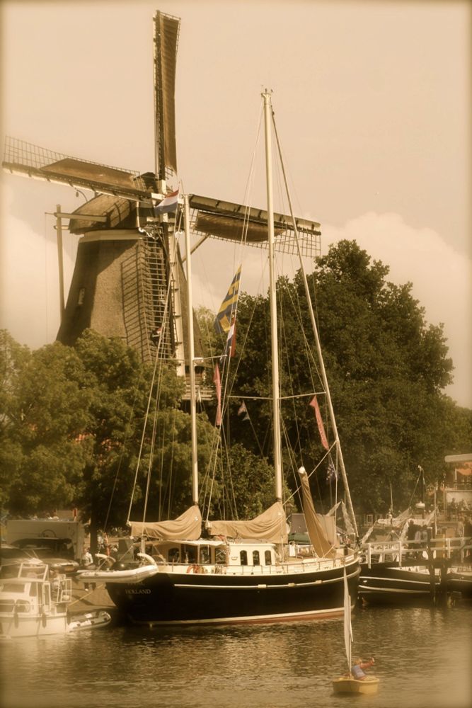 A sepia photo of a windmill surrounded by tall trees. Just in front of it, a boat with tall masts lies in the water. There's another small (slightly blurry) boat with a person at the very bottom of the photo.
