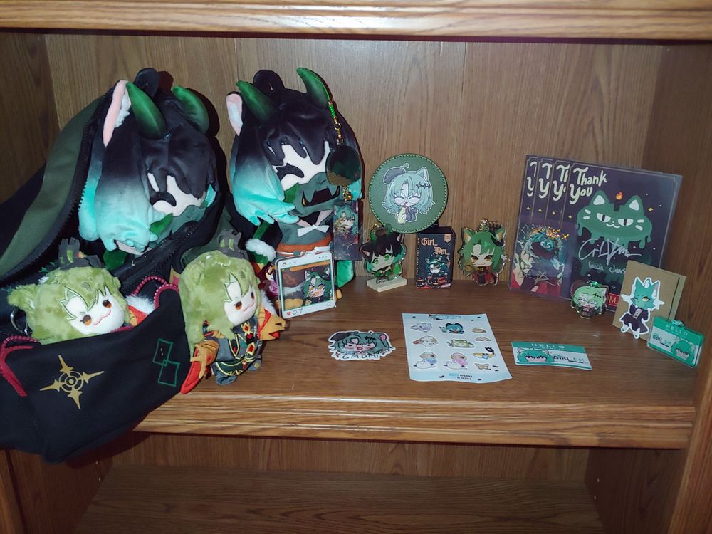 Girl_DM_ merch shelf full view. Left is cross body bag, 2 nui, 2 oni plush, overlay, charm, coaster, keycap, keychain, holographic card, thank you cards from Ohmonah, gremlin sticker, other OffKai merch.