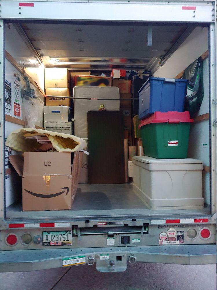 Showing rear of truck. Packed a lot of boxes still from floor to ceiling, have some rope barriers as well as a box spring, with Girl_DM_ nui sitting on edge of a table. Packing material in the front section.