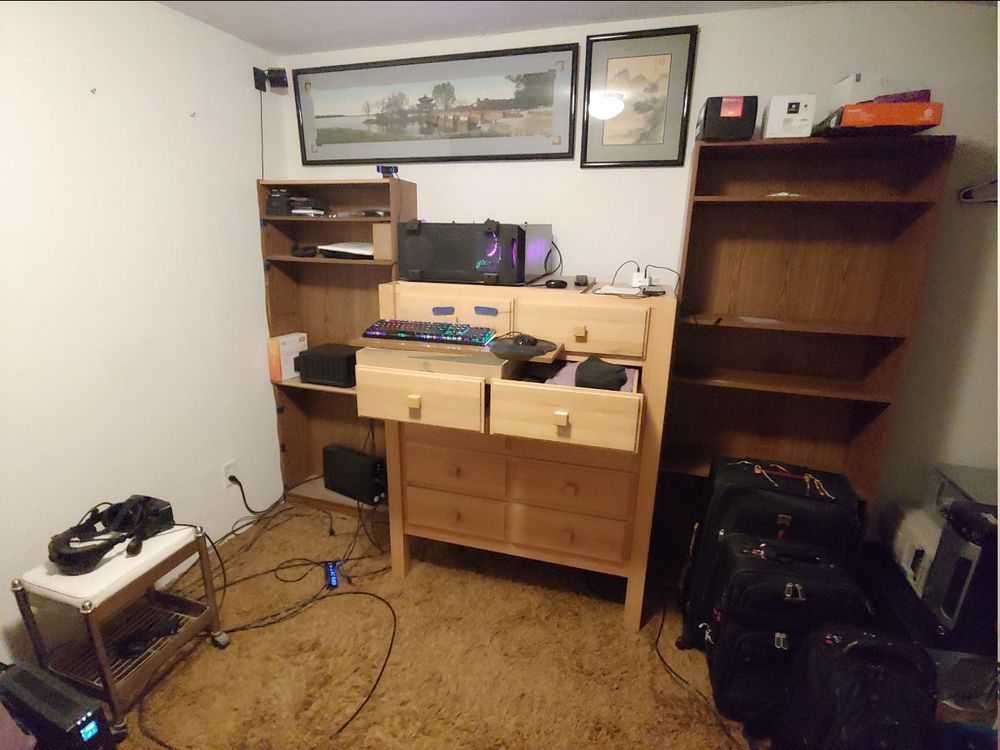 Valve index VR headset sitting on rolling cart to the left. Empty bookcases sit on either side of a tall dresser in the center, with a desktop on its side sitting on top of the dresser. Keyboard and mouse are on a board on the dresser drawers. Suitcases and old computers on the right. Framed pictures above on the wall.