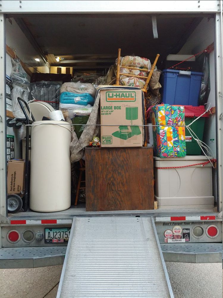 Some dressers and desks, but with a lot of random crates, totes, boxes, chairs, and other odd shaped stuff stacked up and around. Girl_DM_ nui centered on the desk. Showing the back of the uhaul with the ramp extended. Ropes and wires used to tie it all in place.