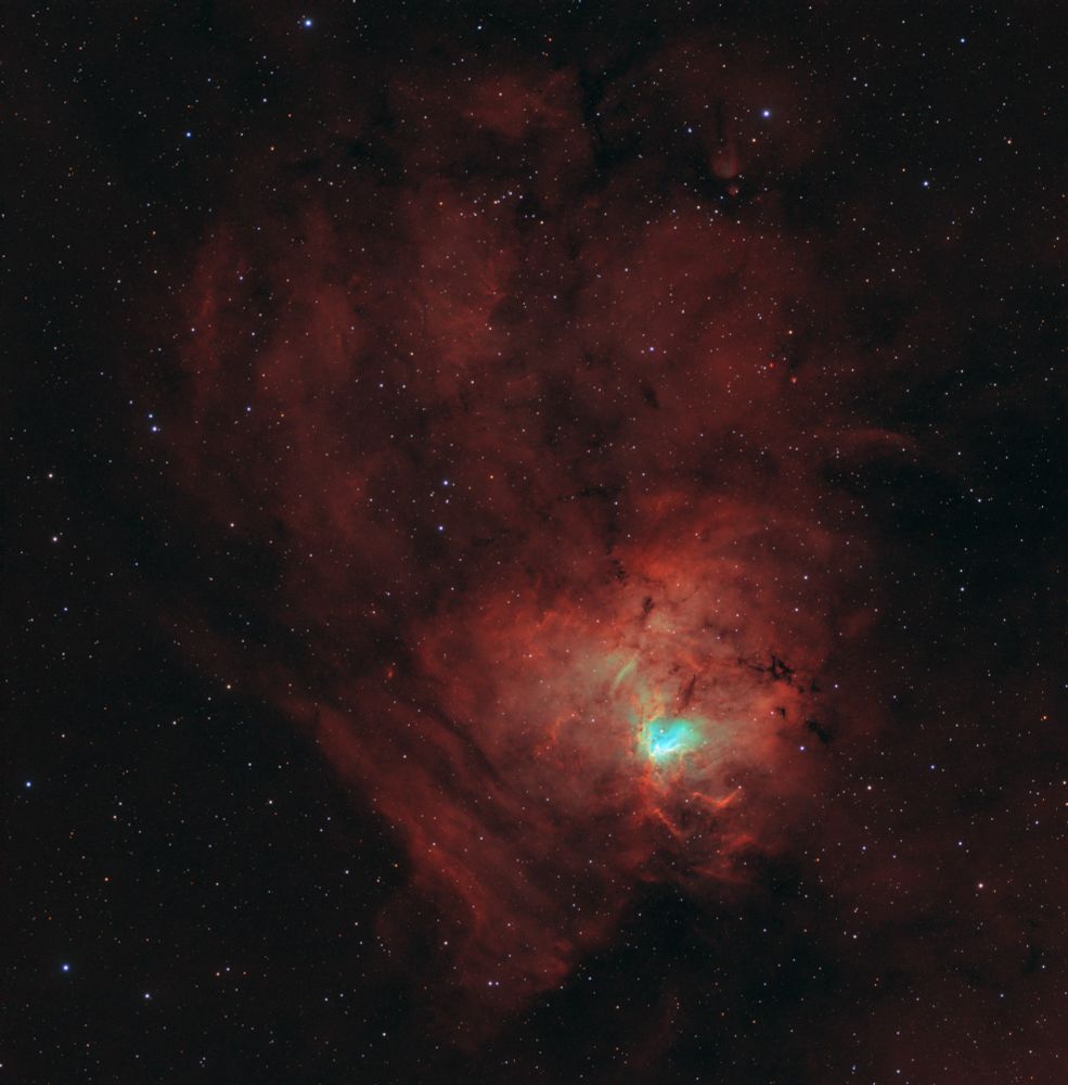 A false color space nebula in reds, oranges and cyans. It’s shaped roughly like a dinosaur footprint. There’s a bright center with complex nebulosities.