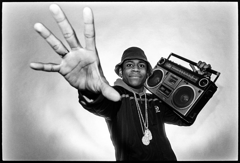 B&W photograph of LL Cool J wearing a bucket hat holding a ghetto blaster on his shoulder, with his hand stretched out to the camera. 